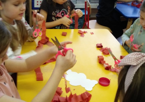 Dzieci siedzą przy stoliku I wykonują pracę plastyczną z bibuly