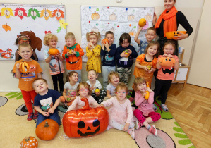 Dzieci stoją z nauczycielką pod tablicą okolicznościową w dyniowych przebraniach