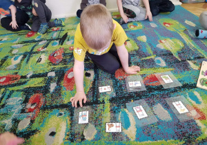 Chłopiec na dywanie przygląda się obrazkom dinozaurów. 