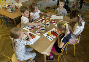 Dzieci siedzą przy stole i malują farbami