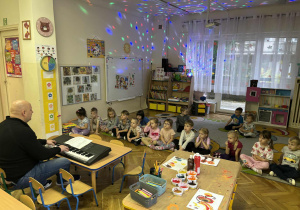 Dzieci siedzą na dywanie i śpiewają piosenkę z prowadzącym