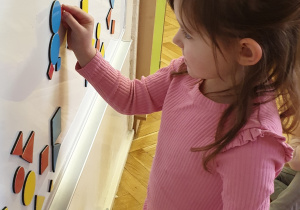 Dziewczynka stoi przy tablicy z figurami geometrycznymi