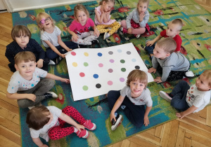 Zdjęcie z góry. Dzieci siedzą na dywanie z kartonem w kropki