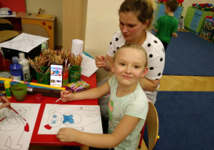 Dziewczynka siedzi przy stole i maluje obrazek farbami