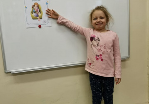 Dziewczynka stoi przy tablicy i wskazuje na obrazek
